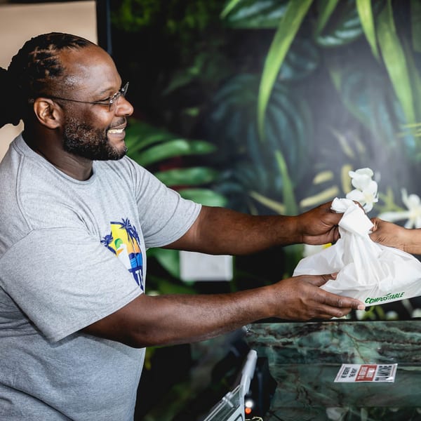A smiling man in glasses hands a compostable takeout bag to a customer at Retreats.