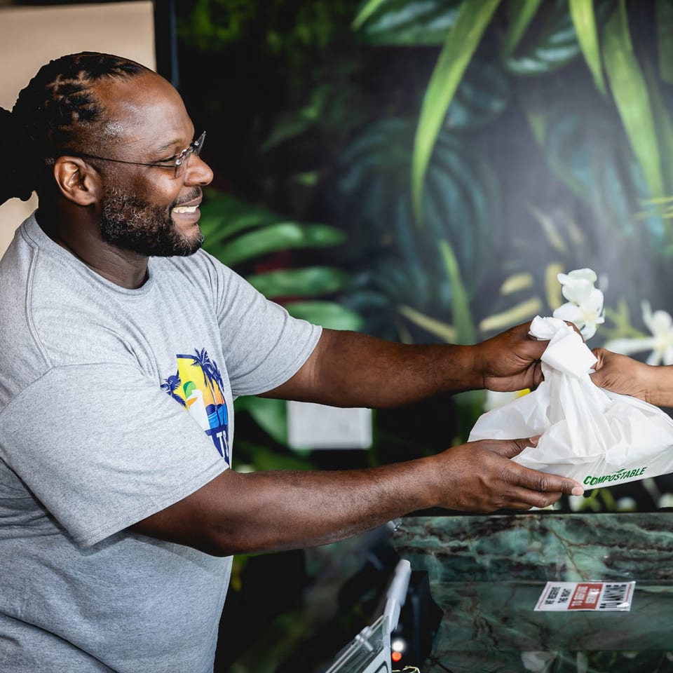 A smiling man in glasses hands a compostable takeout bag to a customer at Retreats.