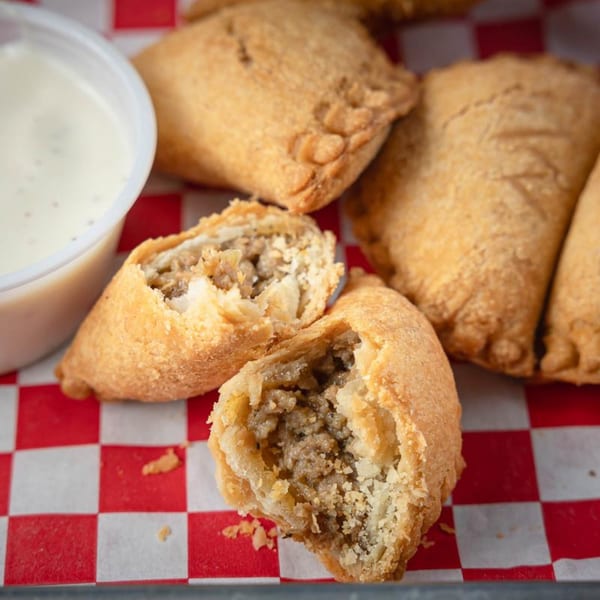 Golden-brown Cajun meat pies, one split open to show the savory filling, with a side of dipping sauce.
