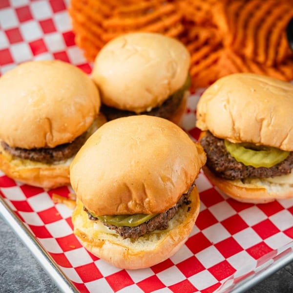 Four sliders with pickles and cheese on a red and white checkered paper with waffle fries.