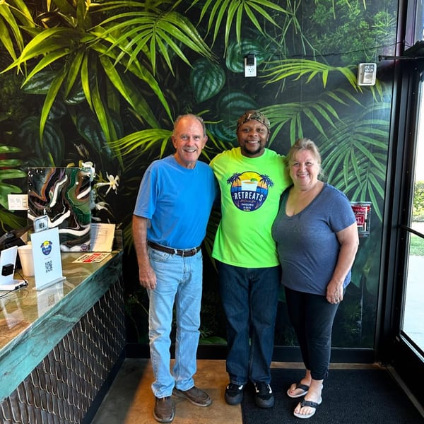 Three smiling people, two customers and one employee in a Retreats shirt, stand in front of a tropical leaf wall.