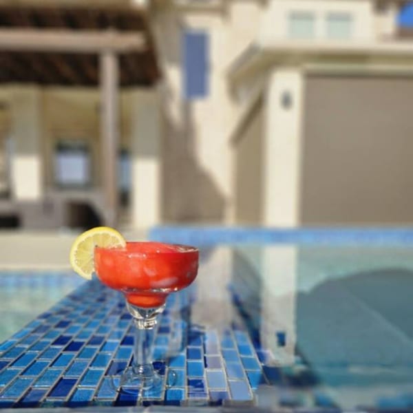 A red daiquiri with a lemon slice sits on blue tiles beside a pool with a building in the background.