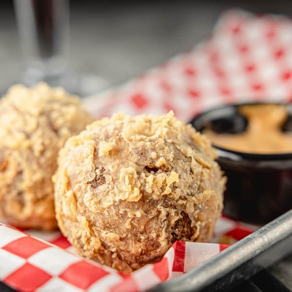Two golden-brown fried boudin balls on red and white checkered paper with dipping sauce.
